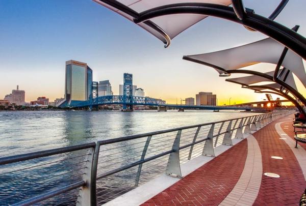 Photo of Jacksonville's Southbank Riverwalk at sunset. Red brick sidewalk alongside the river with steel guardrails. Canopies overhang the sidewalks. Jacksonville skyline in background.